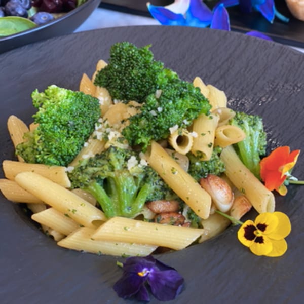 Vegan penne pasta with broccoli, garlic, and edible flowers on a dark plate.