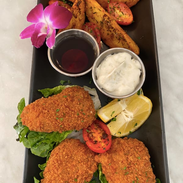 Vegan fish and chips with dipping sauces, lemon, tomatoes, and an orchid on a black platter.