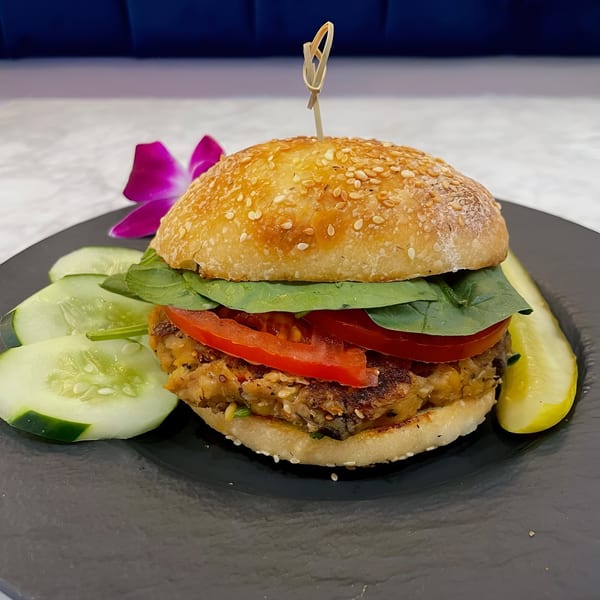 Vegan burger with spinach, tomato, and a sesame bun, served with cucumber slices and a pickle.