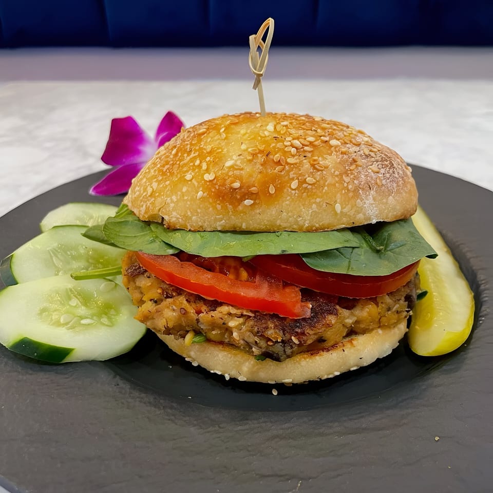 Vegan burger with spinach, tomato, and a sesame bun, served with cucumber slices and a pickle.