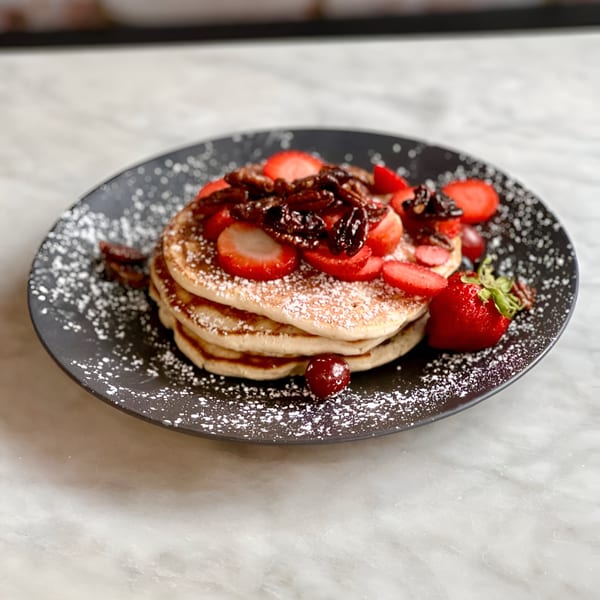 Vegan pancakes with strawberries, candied pecans, and powdered sugar on a dark plate.