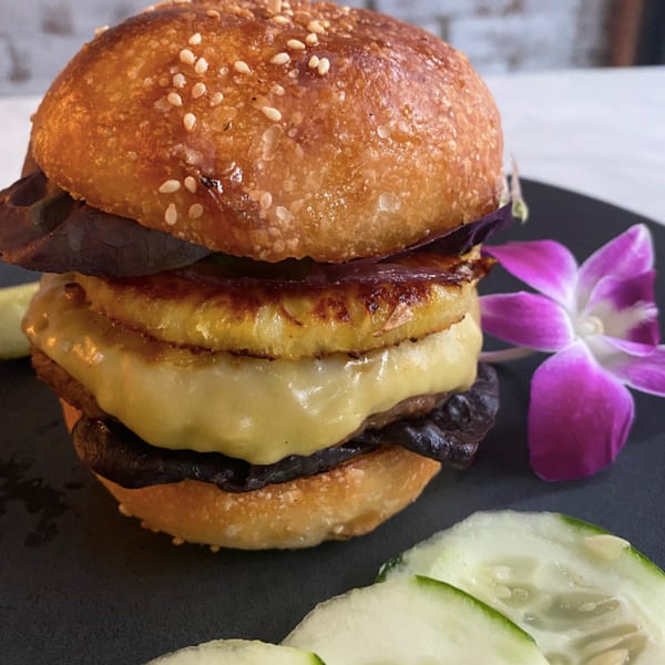 Vegan burger with grilled pineapple, melted cheese, and dark greens, garnished with a purple orchid.